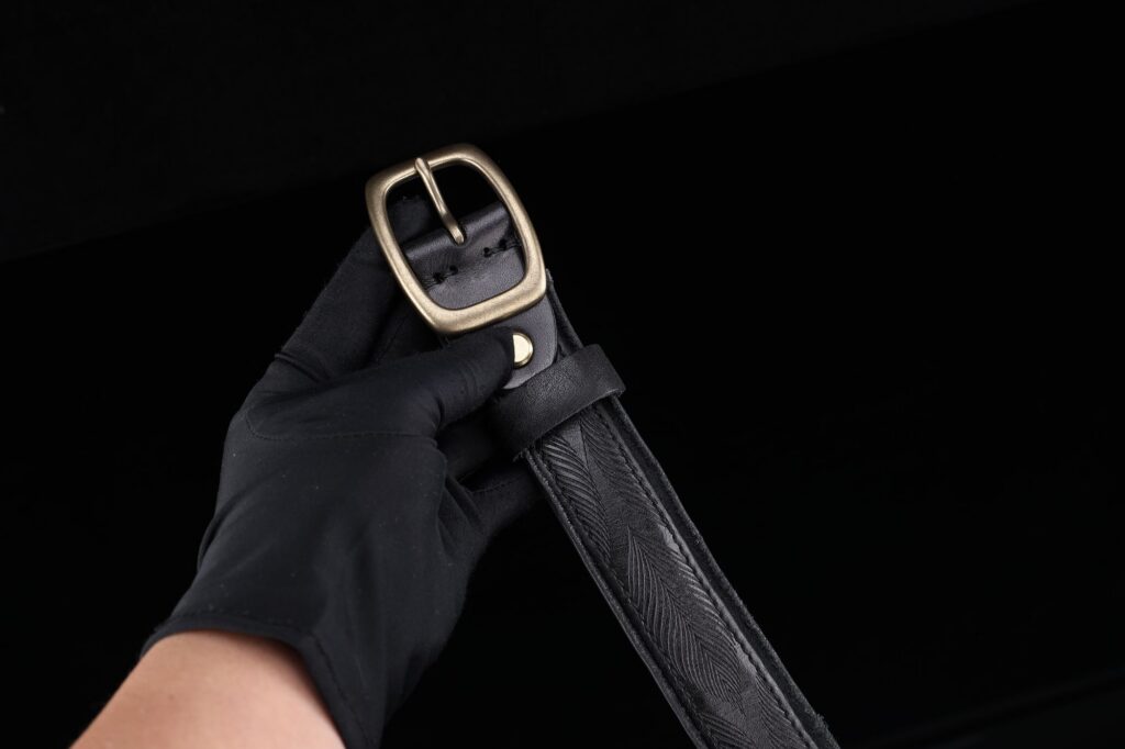 A brass buckle on a black leather belt, held in a gloved hand